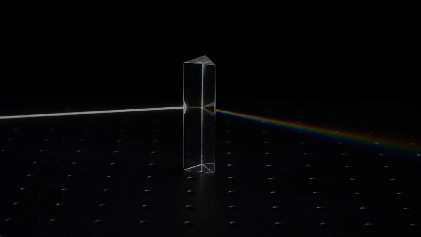 A glass prism on a dark optical breadboard splitting one collimated beam into a spectrum